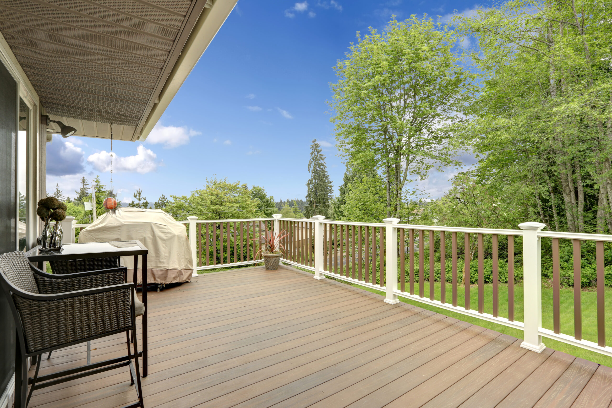 Wooden deck with white and brown railings. Patio table with wicker chairs and barbeque
